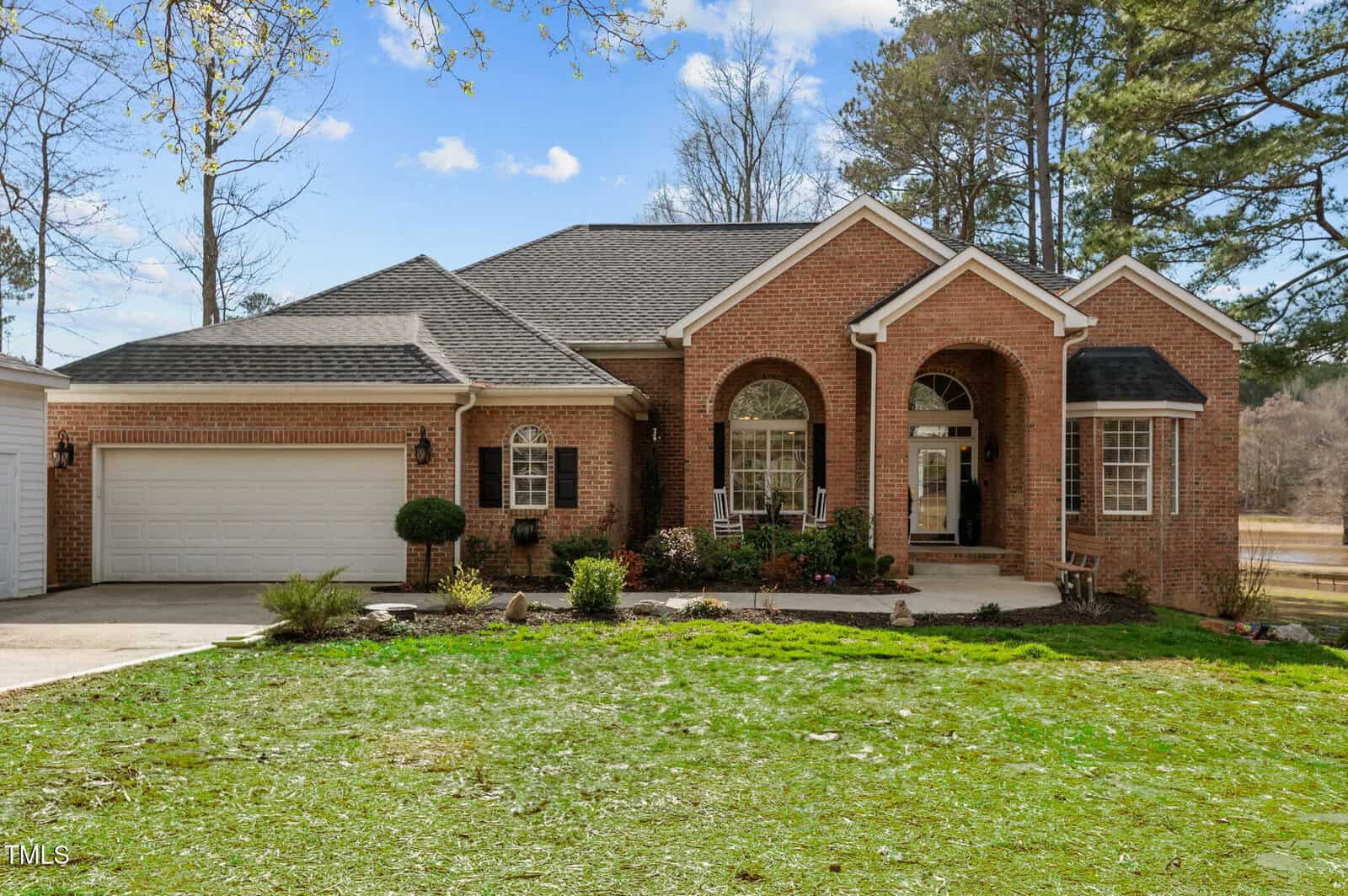 A single-story brick house with arched entryway, white trim, two large front windows, a two-car garage, and a covered front porch. The yard is landscaped with bushes and a green lawn, with trees in the background.