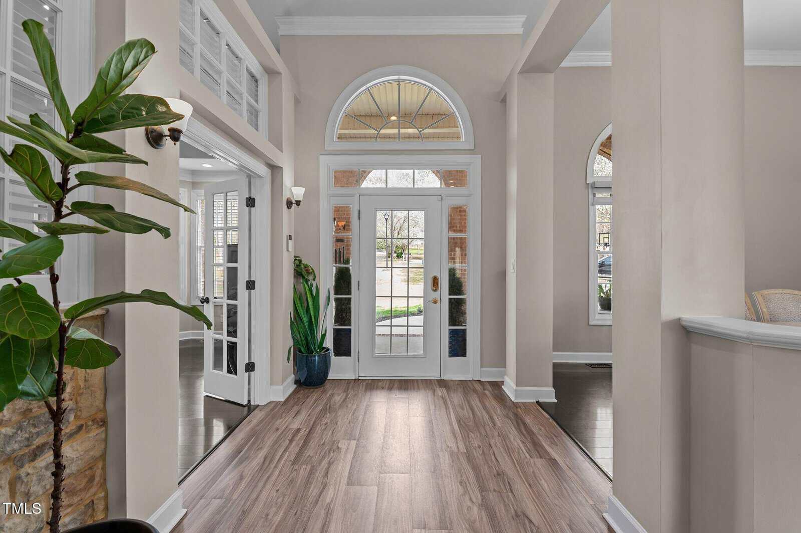 A bright entryway with light wood flooring, cream walls, tall potted plants, and large windows surrounding a glass-paned front door. Outdoor greenery is visible through the windows.