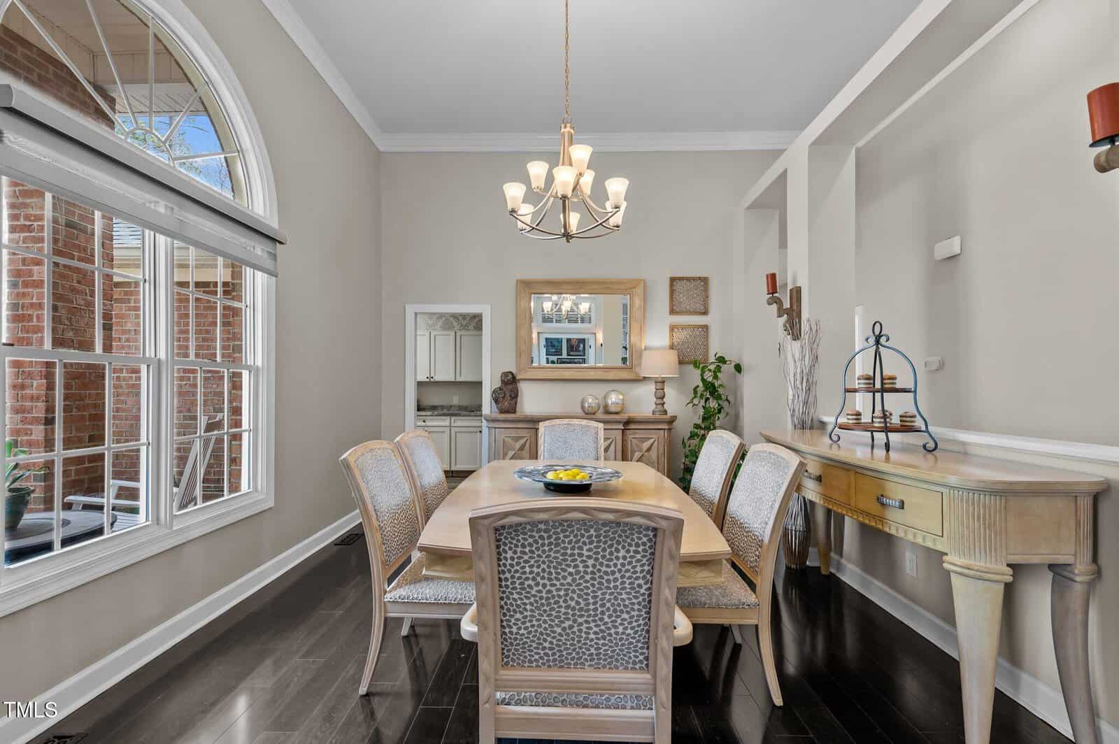 A dining room with a rectangular wooden table and six chairs, a chandelier above, light-colored walls, large window, sideboard with decor, wall niche, and dark wood flooring.