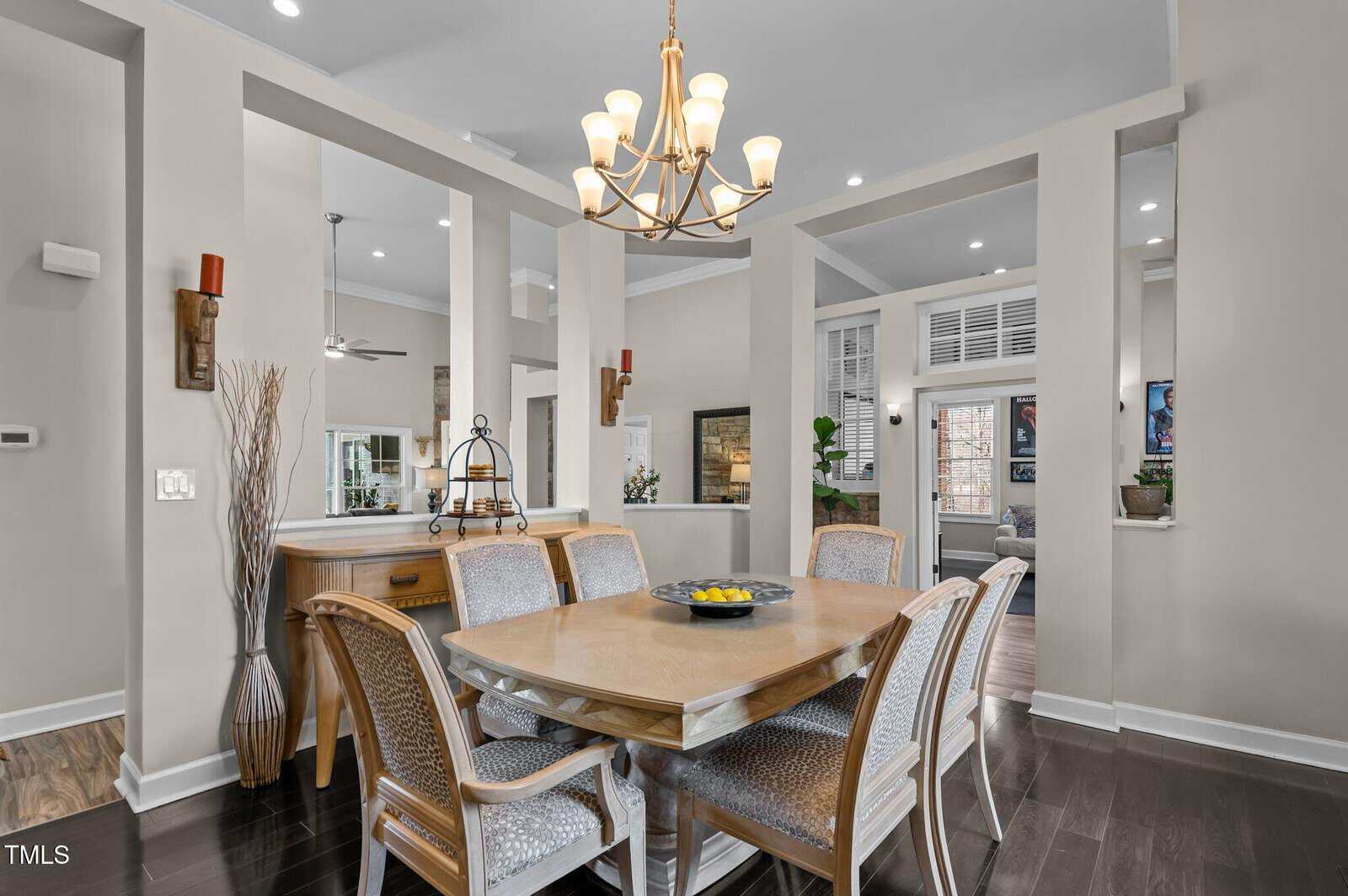 A dining area with a wooden table and six chairs, a chandelier overhead, and a sideboard against a beige wall. Open partitions separate the dining space from adjacent rooms with visible decor and furnishings.