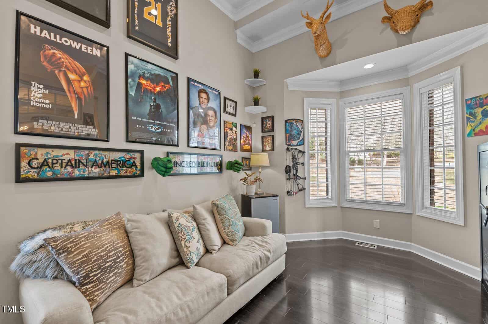 A living room features a beige couch with pillows, movie posters on the wall, two faux deer heads above a window with white shutters, and a TV on a small stand near potted plants.
