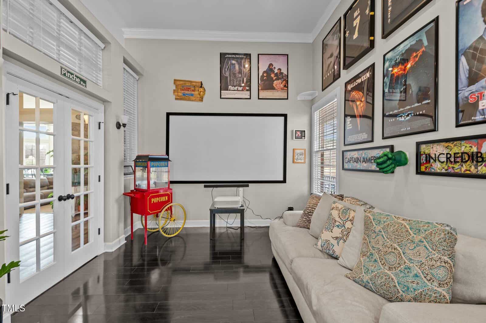Home theater room with a large screen on the wall, movie posters, a popcorn machine, a cushioned beige sofa with assorted pillows, and bright windows with blinds. Hardwood floor and French doors are also visible.