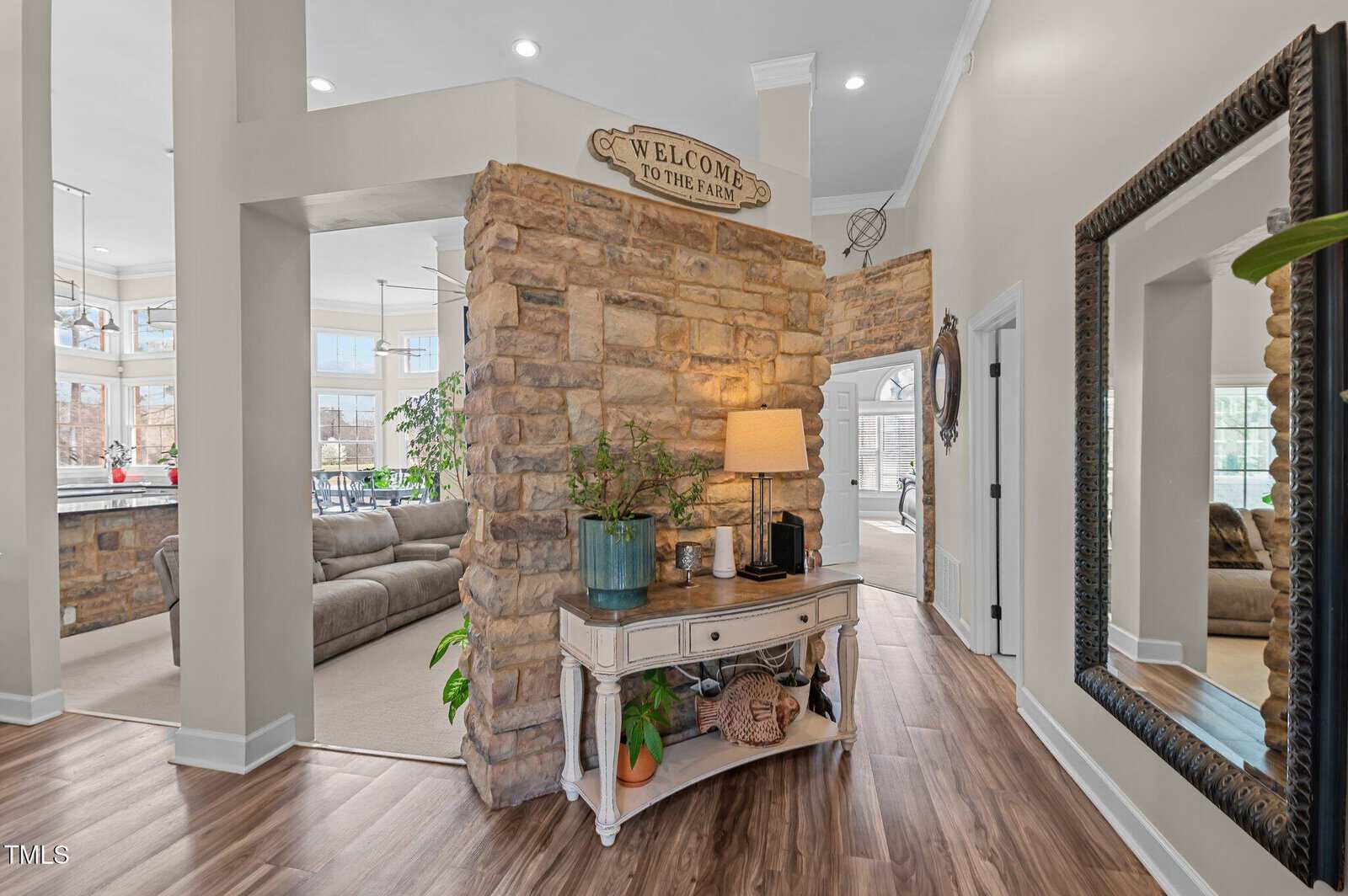 A stone accent wall with a “Welcome to the Farm” sign divides a living area from an entryway with wood flooring, a console table, a lamp, plants, and a large framed mirror on the right wall.
