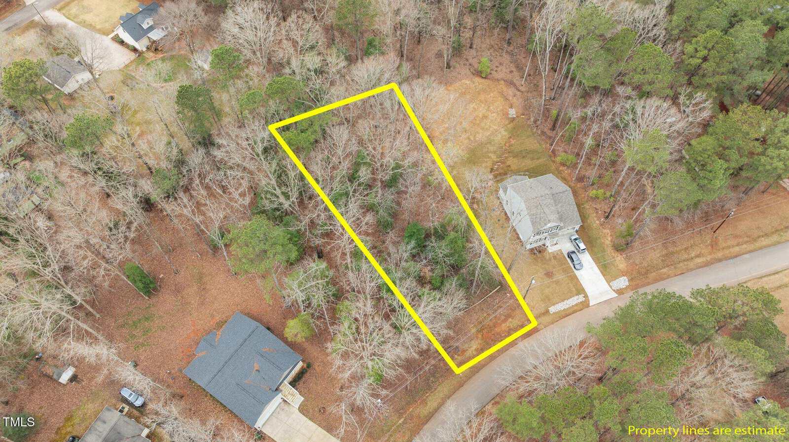 Aerial view of a wooded residential area with a rectangular plot outlined in yellow, showing estimated property lines. Several houses and driveways are visible nearby, and trees surround the highlighted lot.