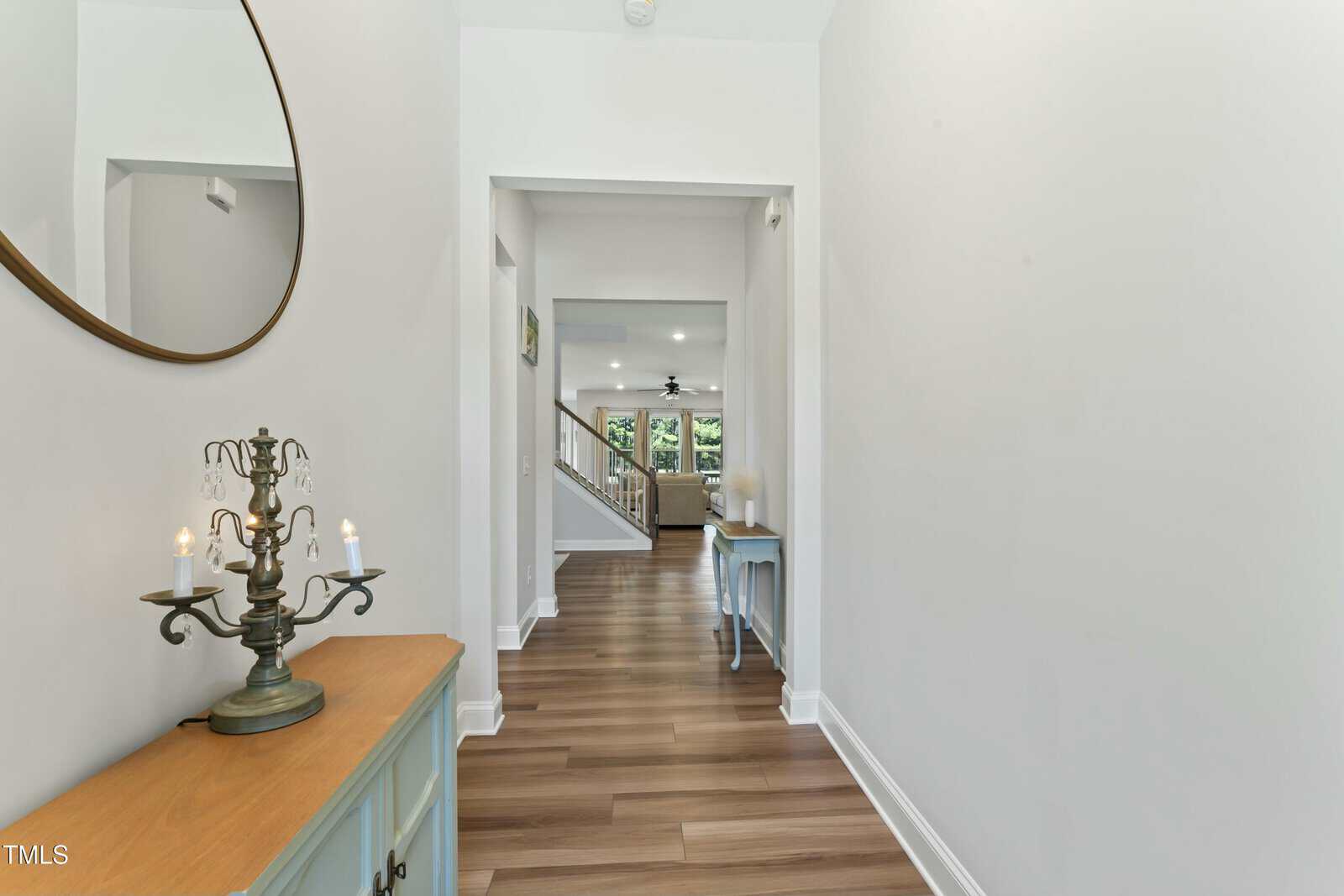 A hallway with light walls and wood flooring leads to a living area. A round mirror, candelabra, and blue console table are on the left. A staircase and windows are visible in the background.