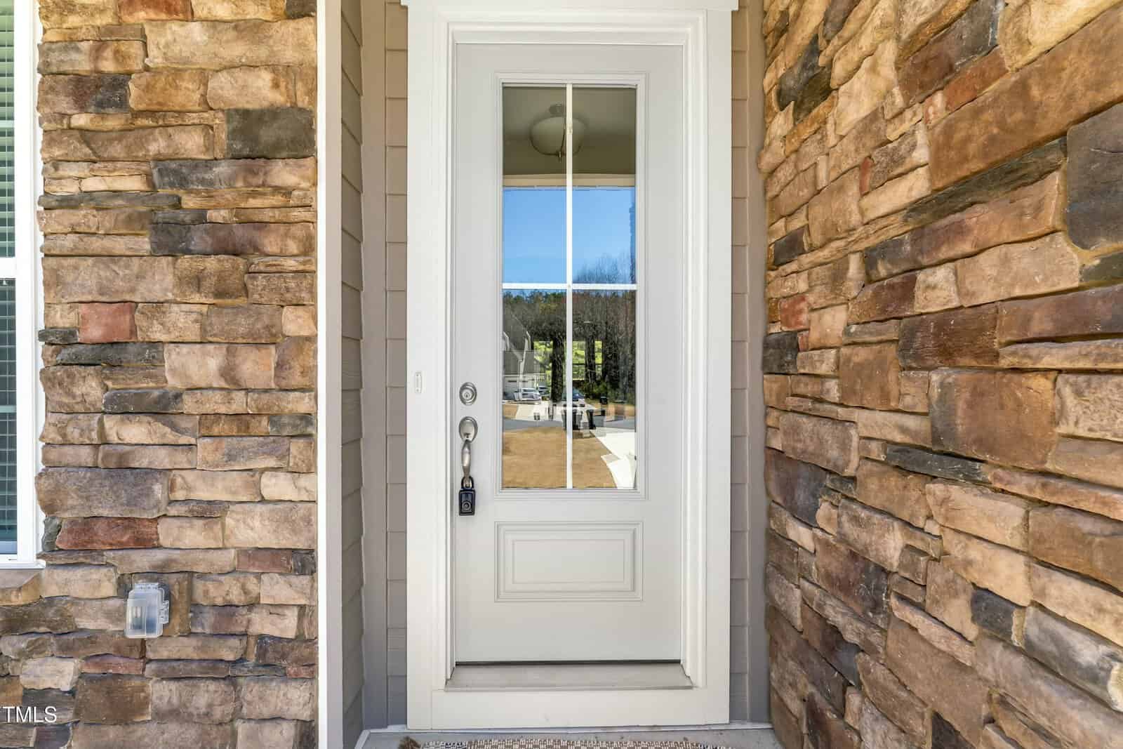 White front door with a glass panel and lockbox, set in a house exterior with brown stone walls. The reflection in the glass shows trees, blue sky, and part of the front yard.
