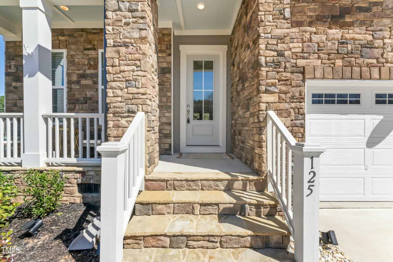 A front entrance with stone steps and a white door, flanked by a white railing and partial view of a garage door with the number 125. The exterior walls are covered with tan and brown stone.