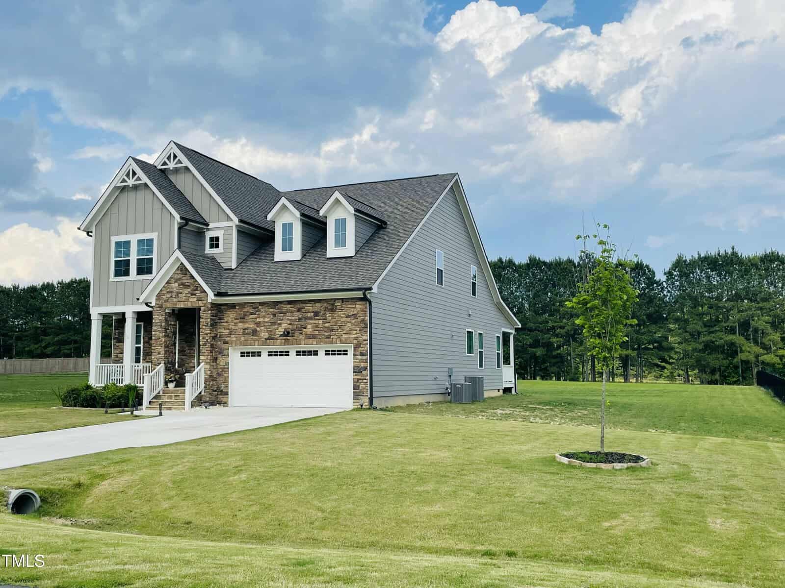 A two-story suburban house with gray siding and stone accents, an attached two-car garage, and a large, well-kept lawn with a young tree. The sky is partly cloudy and there are trees in the background.