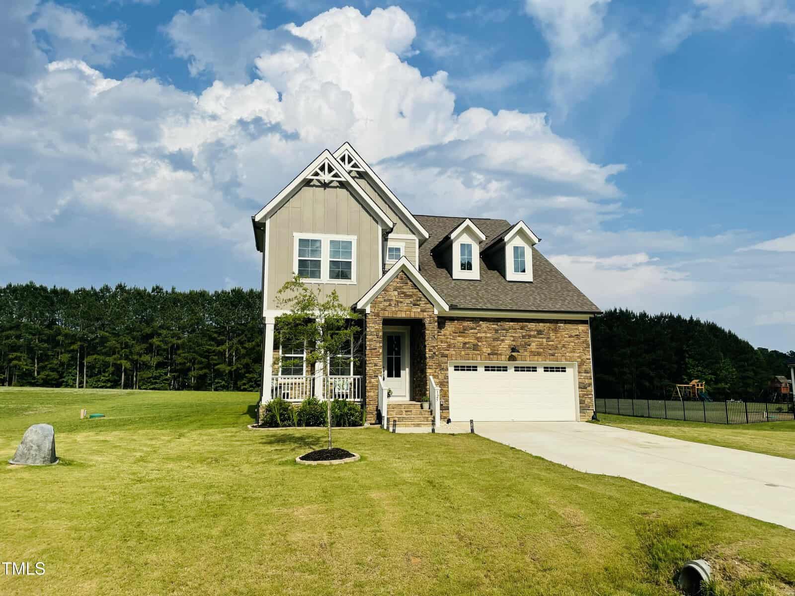A two-story house with a front porch and attached two-car garage sits on a large, neatly trimmed lawn. The house features stone and siding exterior, with a tree planted in the front yard. A wooded area is visible in the background.
