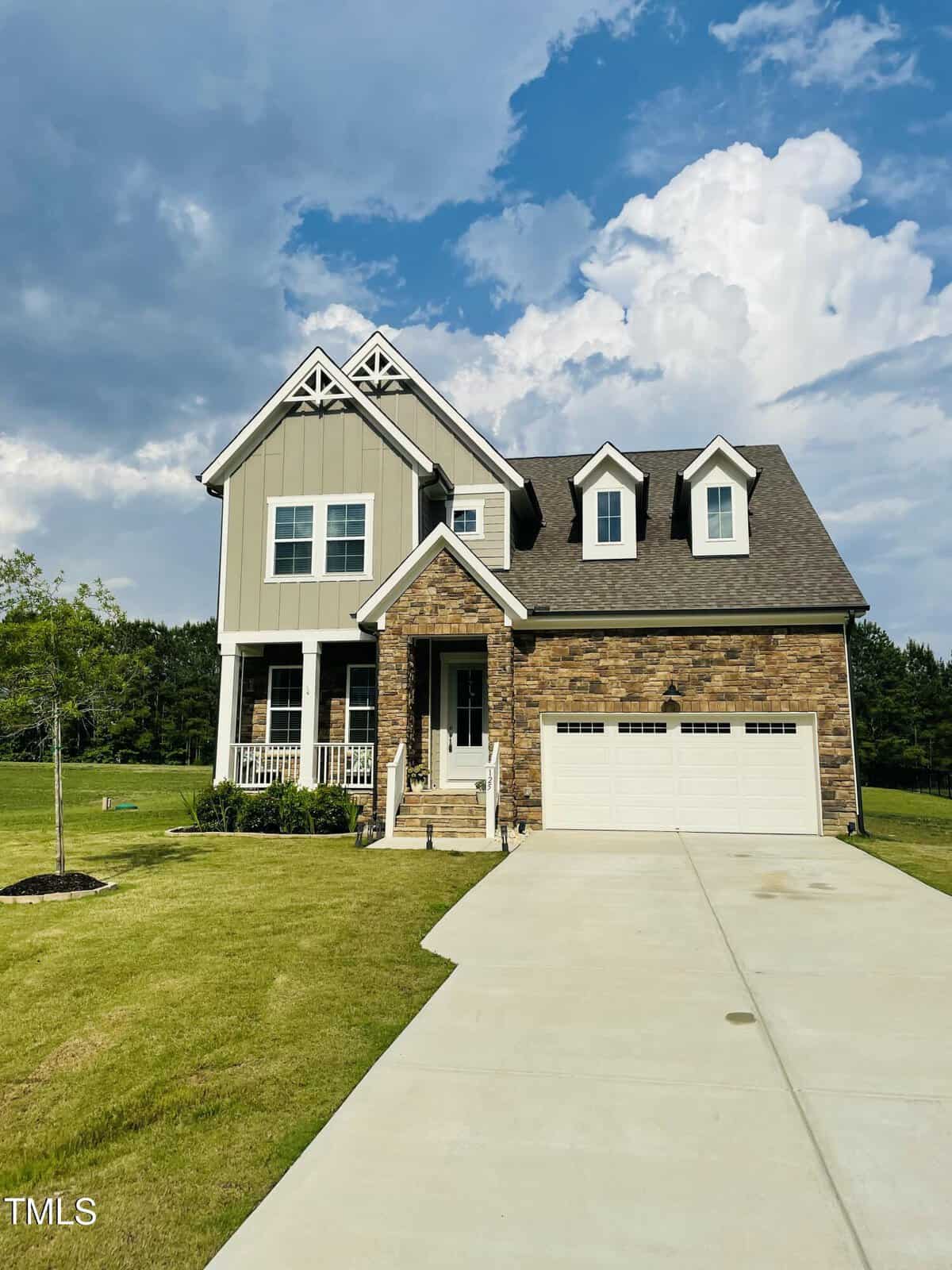 A two-story house with gray siding, stone facade, white trim, and a front porch. The home has a double garage and sits on a lawn with a driveway and young trees, under a partly cloudy sky.