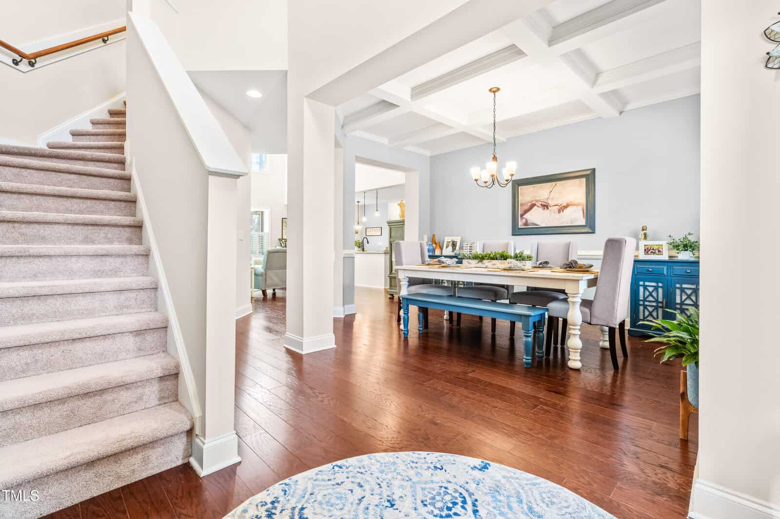 A dining room with a coffered ceiling, chandelier, blue sideboard, and long table set for a meal. Hardwood floors and white walls are visible, with a carpeted staircase to the left and a blue patterned rug in the foreground.