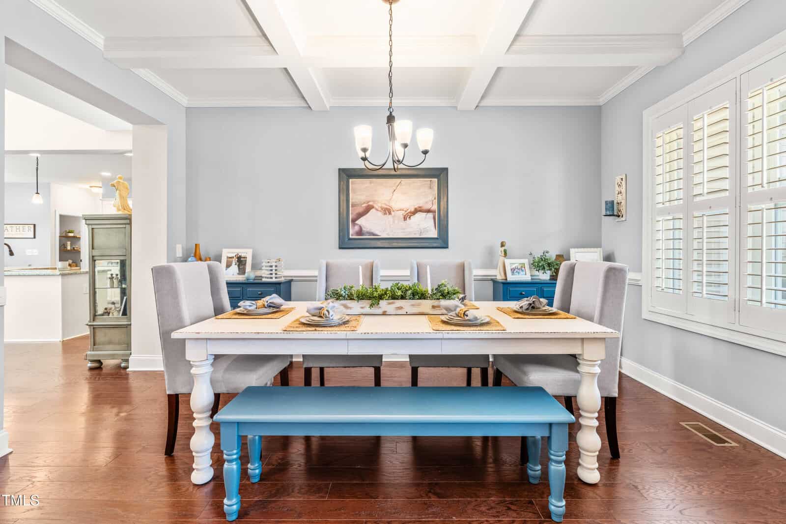 A dining room with a white and blue table, six upholstered chairs, and a blue bench. Table is set for four. A chandelier hangs from the coffered ceiling. Artwork and cabinets are visible along the light blue walls.