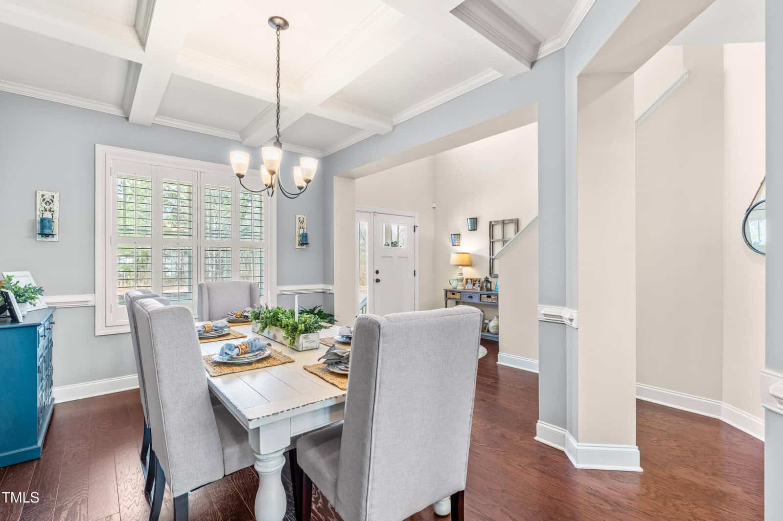 Bright dining room with light gray walls, a white coffered ceiling, wooden floors, a rectangular table set for four, upholstered chairs, a blue sideboard, and large windows with white plantation shutters.