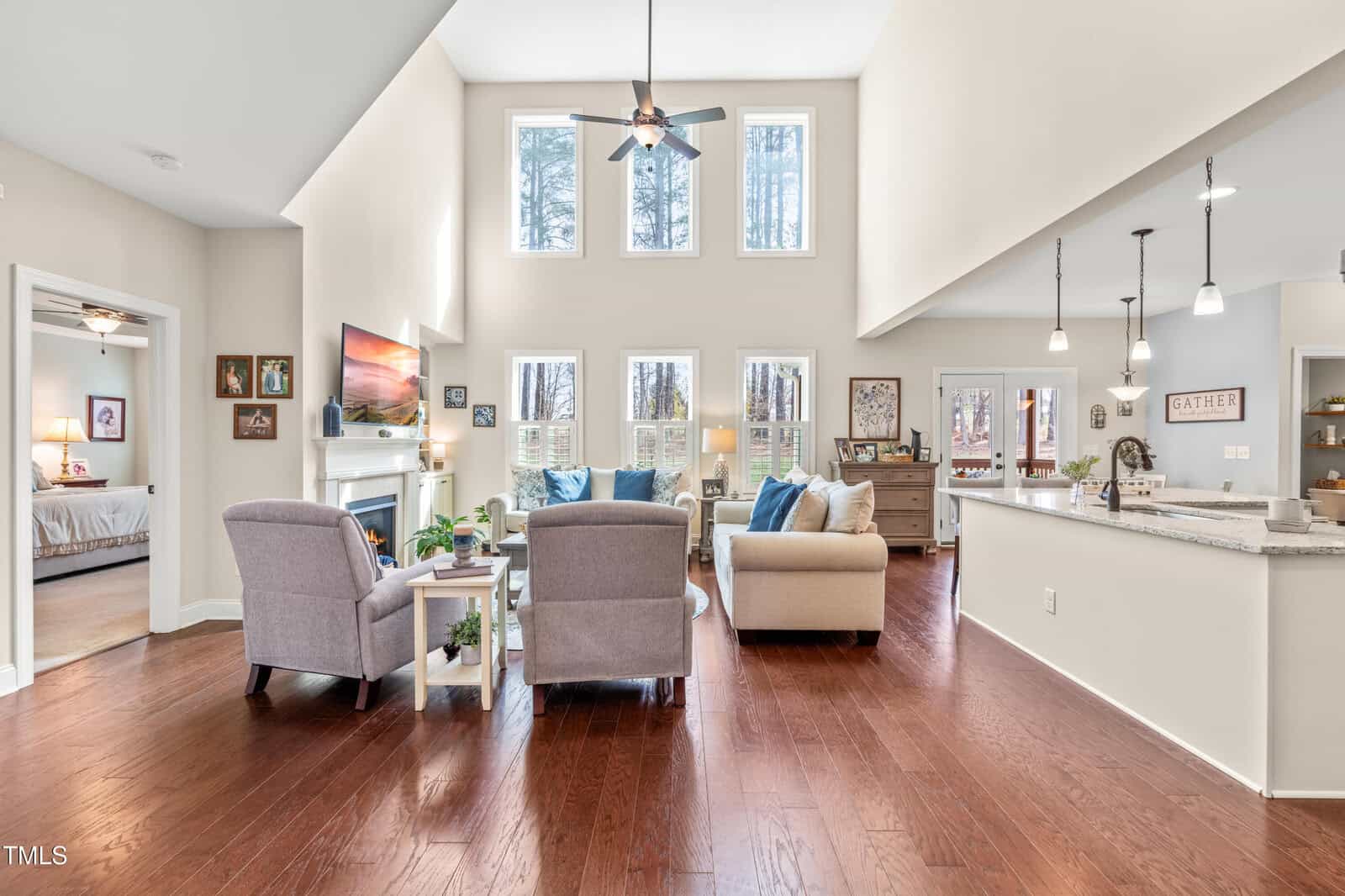 Open-concept living room with high ceilings, large windows, light-colored walls, hardwood floors, and modern furniture. A kitchen with pendant lighting and a breakfast bar is visible to the right, and a bedroom is to the left.