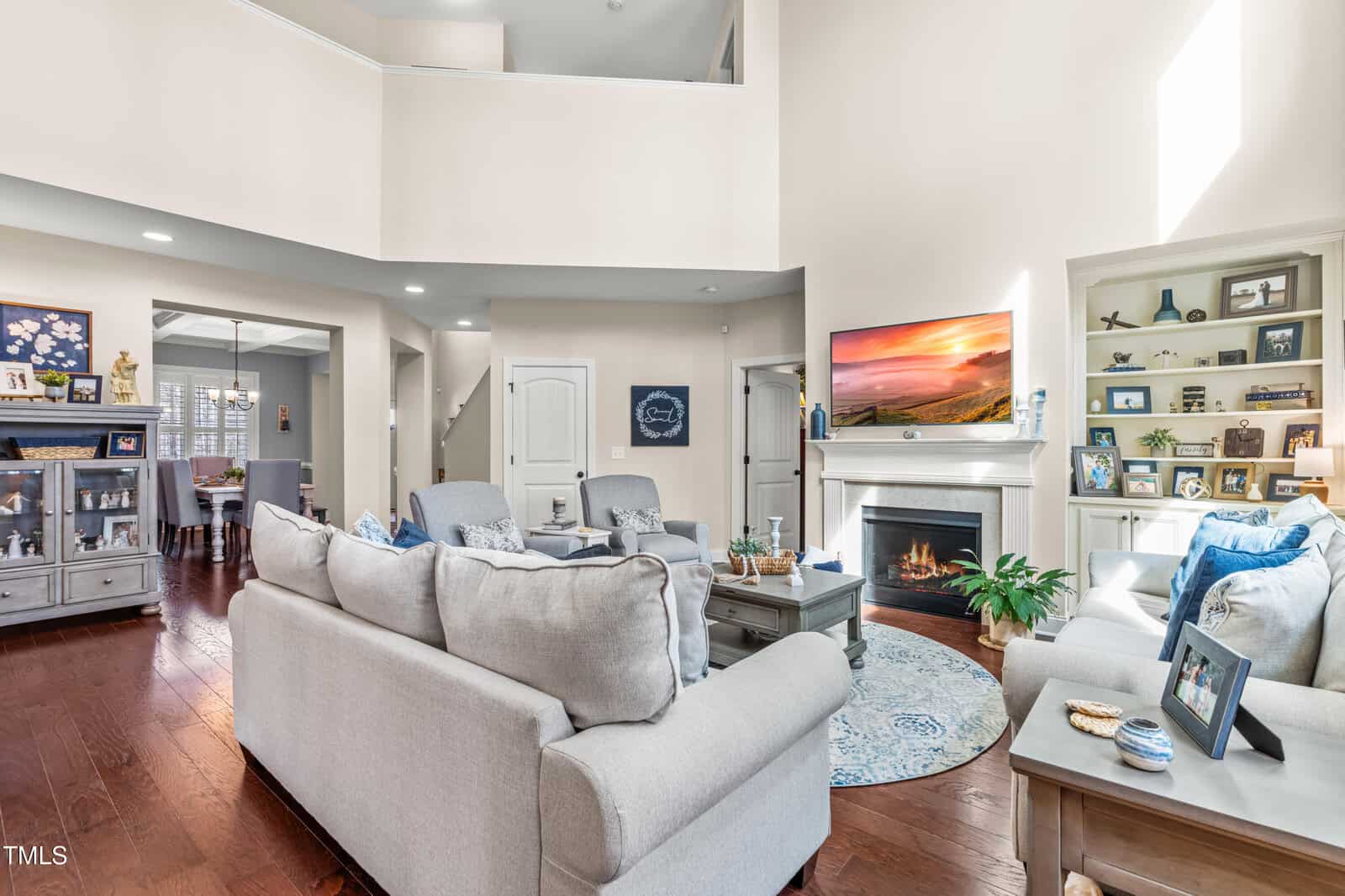 Bright living room with light gray sofas, a coffee table, a lit fireplace with a TV above it, hardwood floors, and built-in shelves with framed photos and decor. Dining area and stairway visible in the background.