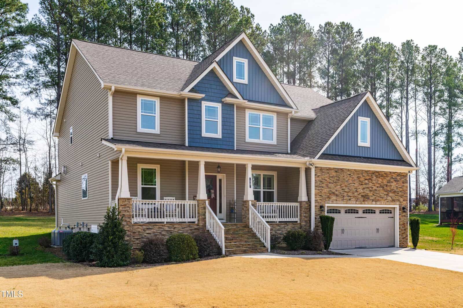 Two-story house with tan siding, blue accents, and stonework details. Covered front porch with white railings, two-car garage, and a neatly trimmed yard. Surrounded by tall pine trees in the background.