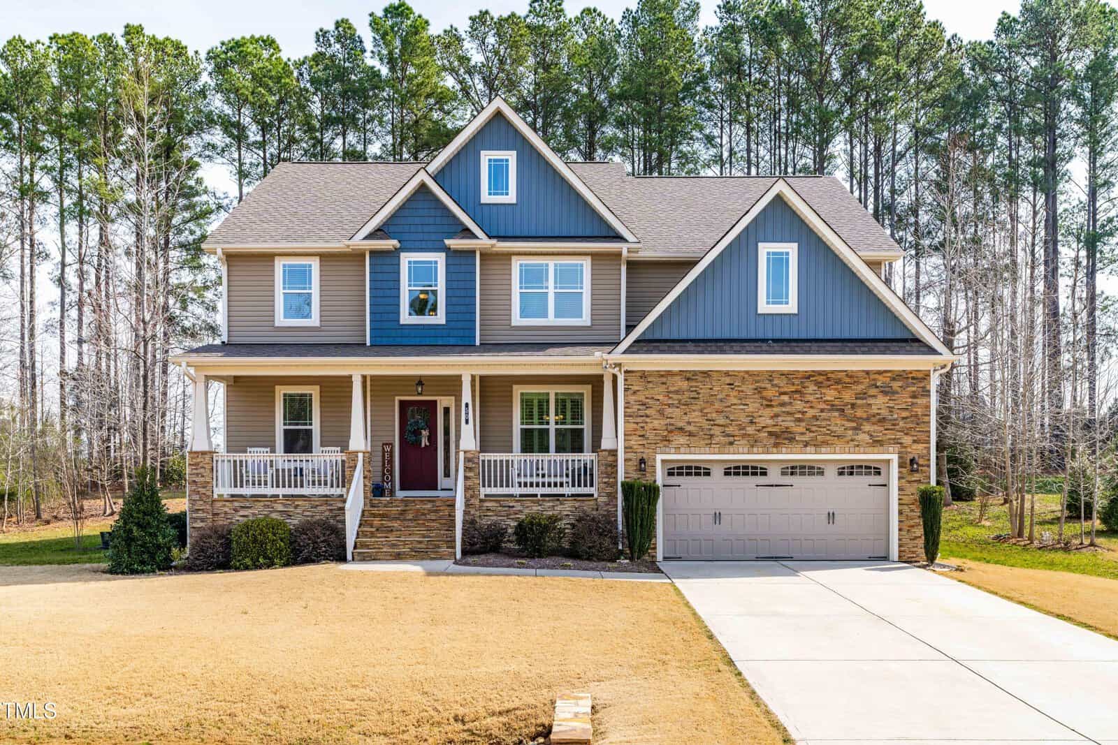 Two-story suburban house with beige siding, blue accents, and stone facade. Features a covered front porch, double garage, and a well-maintained lawn. Tall trees line the background.