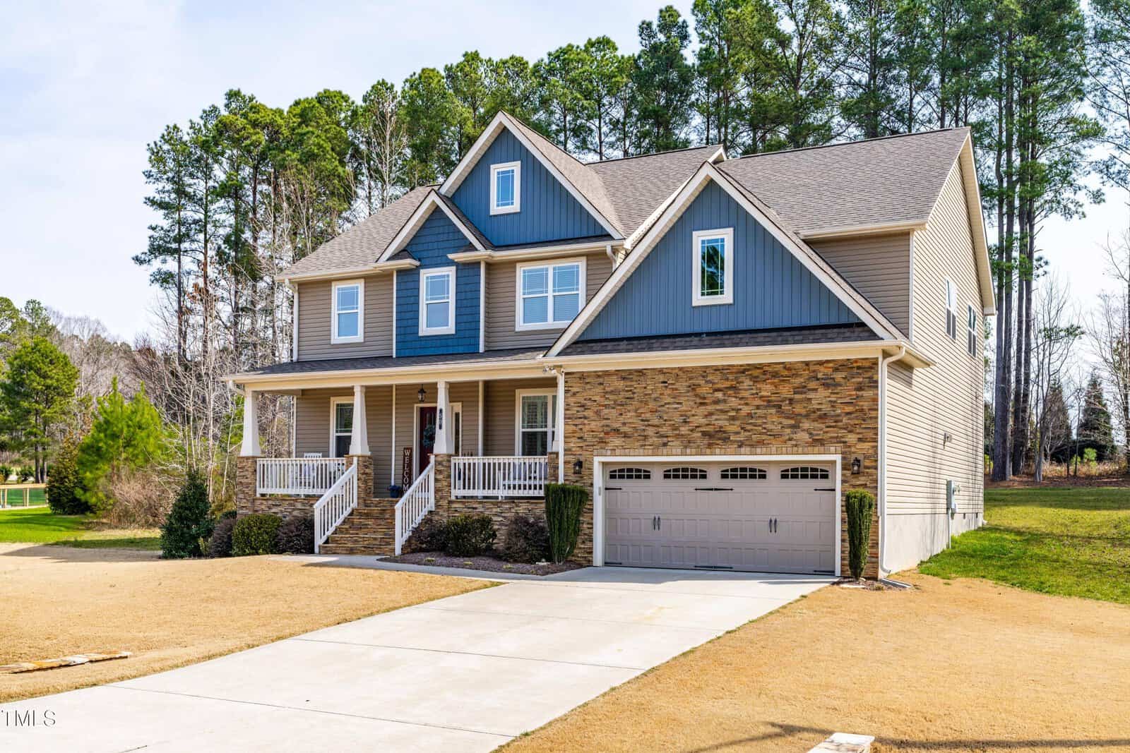 A two-story suburban house with stone and blue siding exterior, a covered front porch, white railings, and a double garage, set on a large lot with trees in the background and a wide driveway in front.