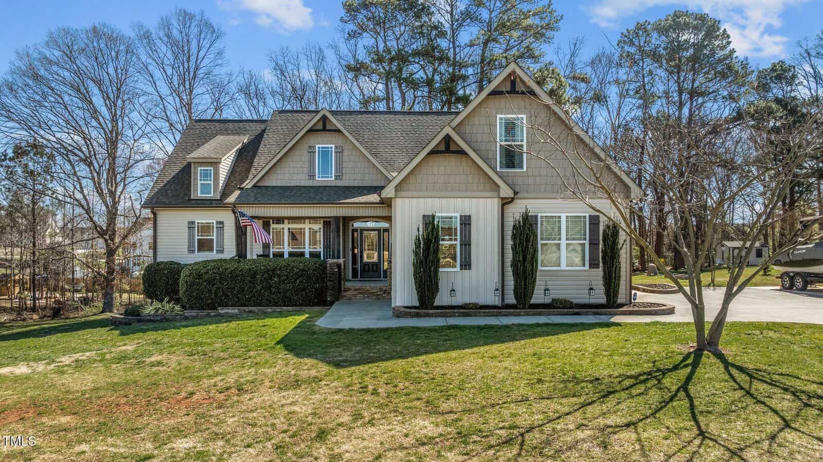 A two-story house on 1046 Lawrence Road in Wake Forest features a gray and beige exterior with a gabled roof. An American flag flutters near the entrance. The grassy yard has sparse trees, and a driveway extends to the right under the clear blue sky.