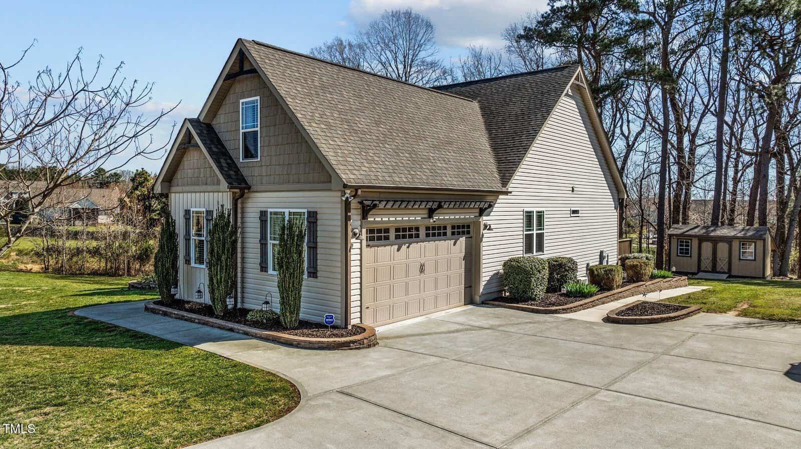 A two-story house with beige siding and a dark roof, featuring a large garage with decorative windows. The front yard is landscaped with shrubs and a well-maintained lawn. A small shed is visible in the background, surrounded by trees.