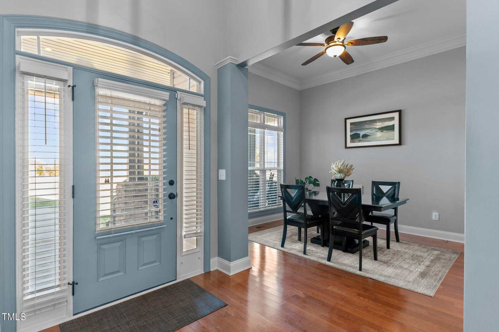 A dining area is visible through an open archway. It features a dark wood table with black chairs on a gray rug, under a ceiling fan. Large windows with blinds and a glass door are on the left, allowing natural light to fill the room.