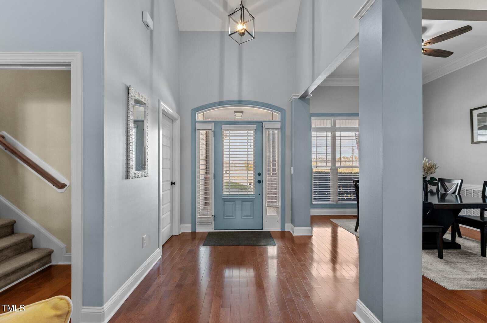 A bright foyer with light blue walls and a wooden floor. A staircase with carpeted steps is on the left. A blue front door with glass panels sits in the center, flanked by windows. A dining area with a dark table is visible on the right.