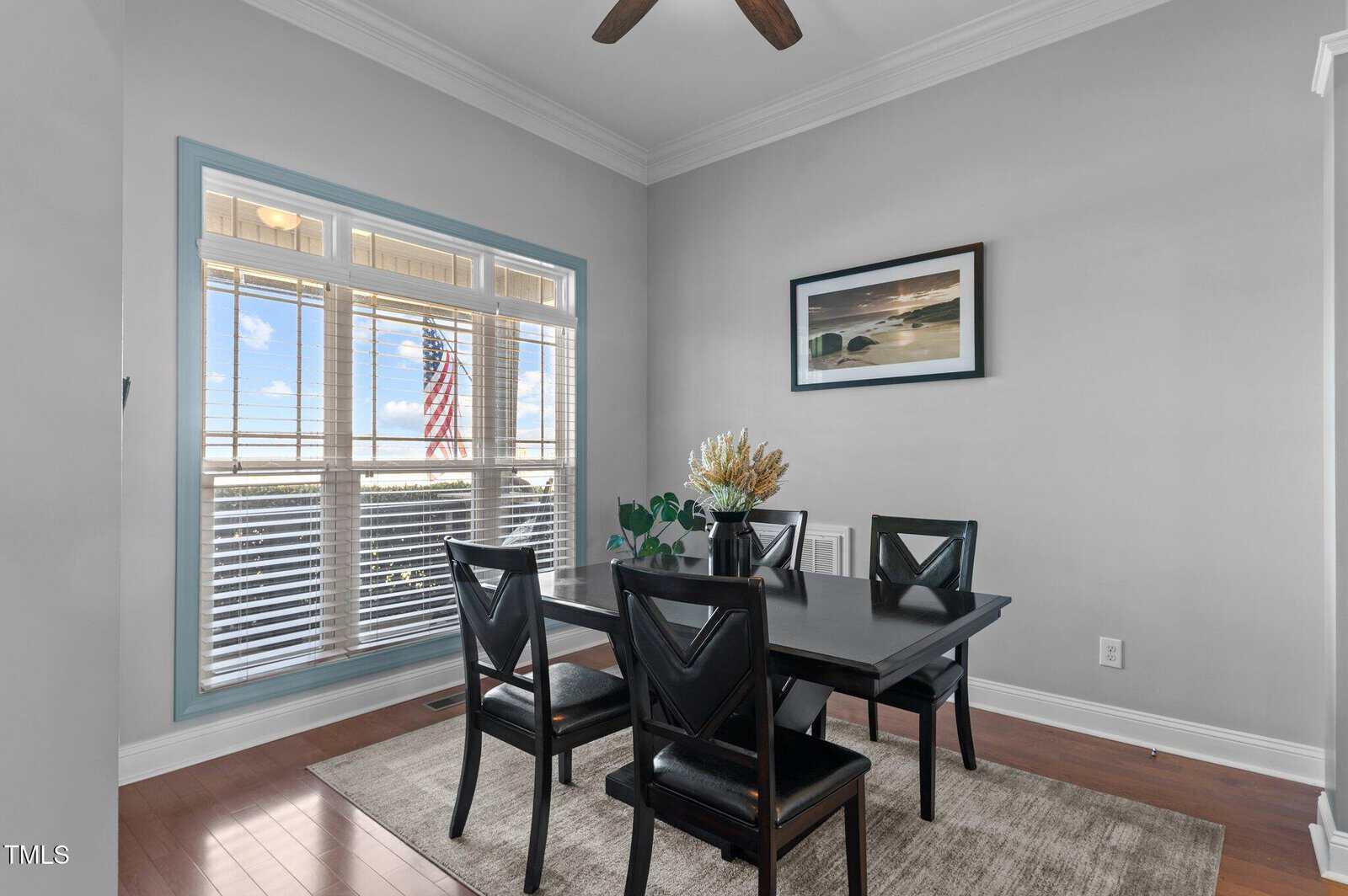 A dining area with a black table and four chairs on a beige rug. A window with blinds lets in natural light and shows a view of an American flag outside. A framed landscape photograph hangs on the light gray wall.