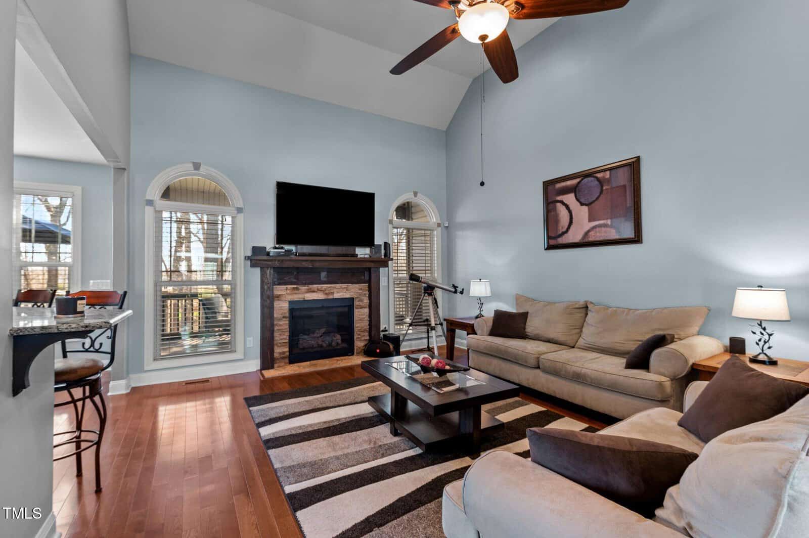 A living room with blue walls and a vaulted ceiling. There's a beige sofa, armchair, and coffee table on a striped rug. A flat-screen TV is mounted above a fireplace. Art decorates the wall. A ceiling fan hangs above, and a window offers outside views.