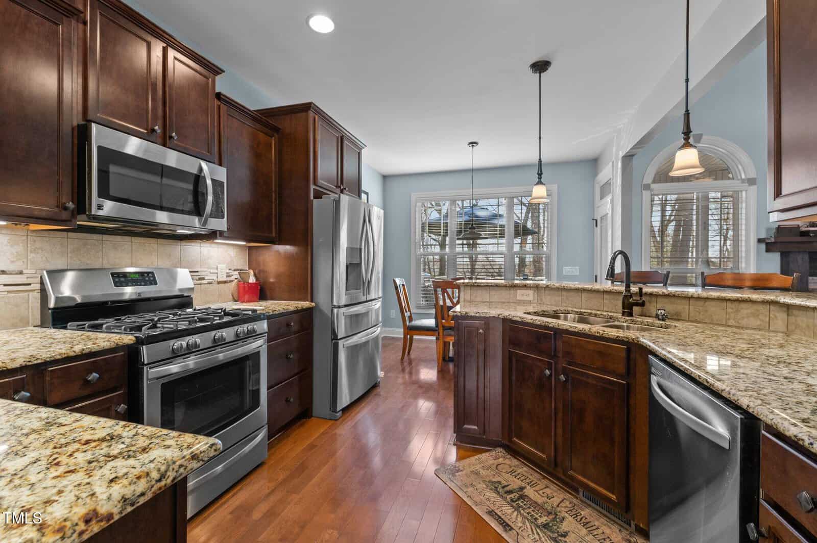 Modern kitchen with dark wood cabinetry and granite countertops. Includes a stainless steel oven, microwave, refrigerator, and dishwasher. A central island with a sink has pendant lighting. A dining table and large window are in the background.