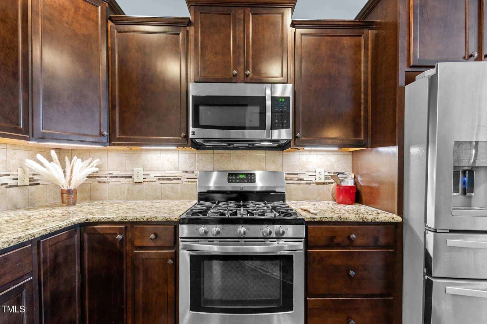 A modern kitchen with dark wood cabinets and stainless steel appliances. A microwave is above a gas stove with an oven. A refrigerator is on the right, and a vase with white flowers is on the granite countertop to the left.
