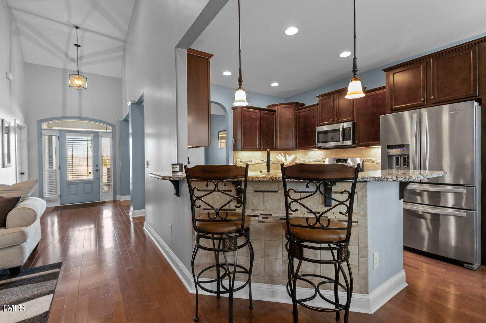 A modern kitchen area with dark wood cabinets, stainless steel appliances, and a granite countertop. Two ornate barstools are at the counter. The space is connected to a hallway with a light blue front door and natural light coming through windows.