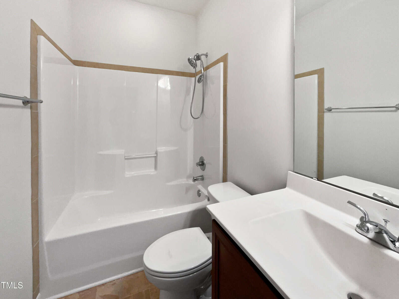 A small bathroom with a white bathtub-shower combo, toilet, and white sink with a chrome faucet. The walls are white, and there is a mounted showerhead above the bathtub. The sink cabinet is dark wood.