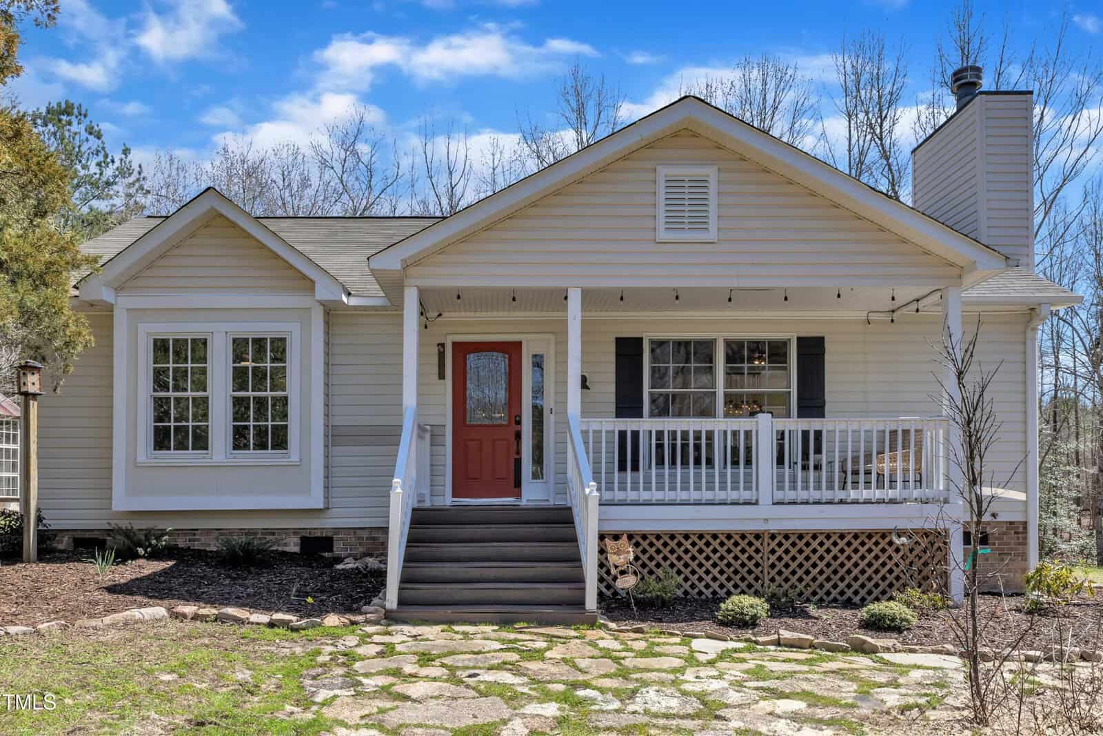 A charming single-story house at 1384 Sims Bridge Road in Kittrell, this home boasts light beige siding and a striking red front door. It features a cozy covered porch with white railings, a right-side chimney, and a short stone path leading up to the inviting entrance under the blue sky.