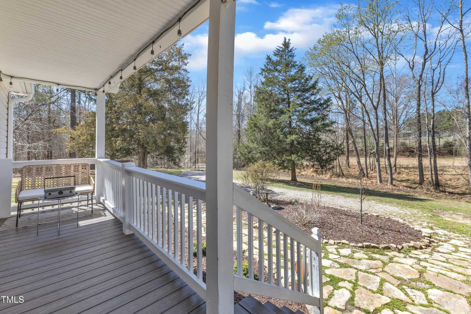 A white wooden porch with a roof, featuring a small table and two chairs, overlooks a stone pathway and a grassy yard. Trees and a clear blue sky are visible in the background.