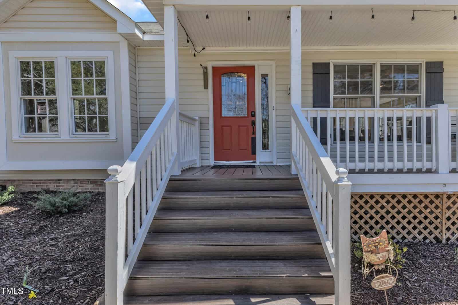 Front view of a house featuring a red door at the top of wooden steps. The porch has white railings and a lattice design underneath. Two large windows are on the left, and a small welcome sign is on the right.