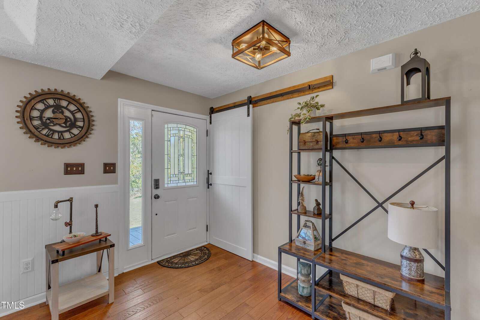 Entryway with a white front door featuring a decorative glass panel. A wooden shelving unit displays plants and decorative items on the right. A rustic clock hangs on the wall. A small table with a lamp is near the door. Wood flooring and a ceiling light.