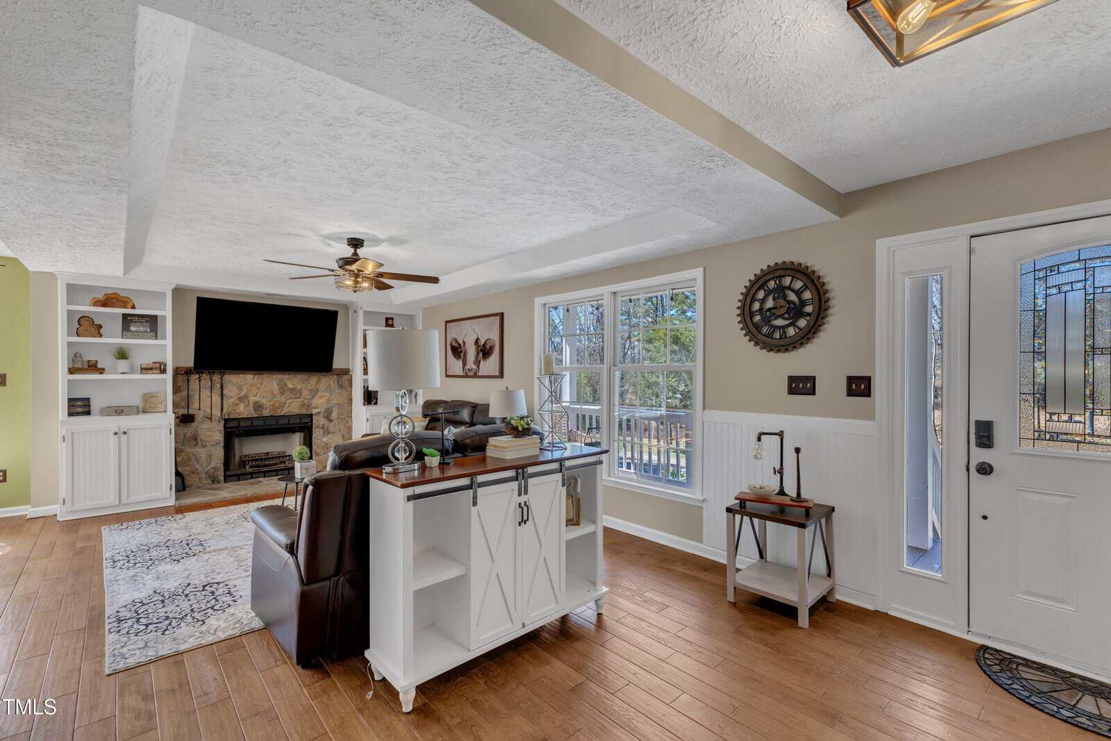 Living room with hardwood floors, white walls, and a textured ceiling. Features a dark sofa, TV on a stone fireplace, ceiling fan, and large windows. A white console table with storage divides the space, next to a white door with glass panels.