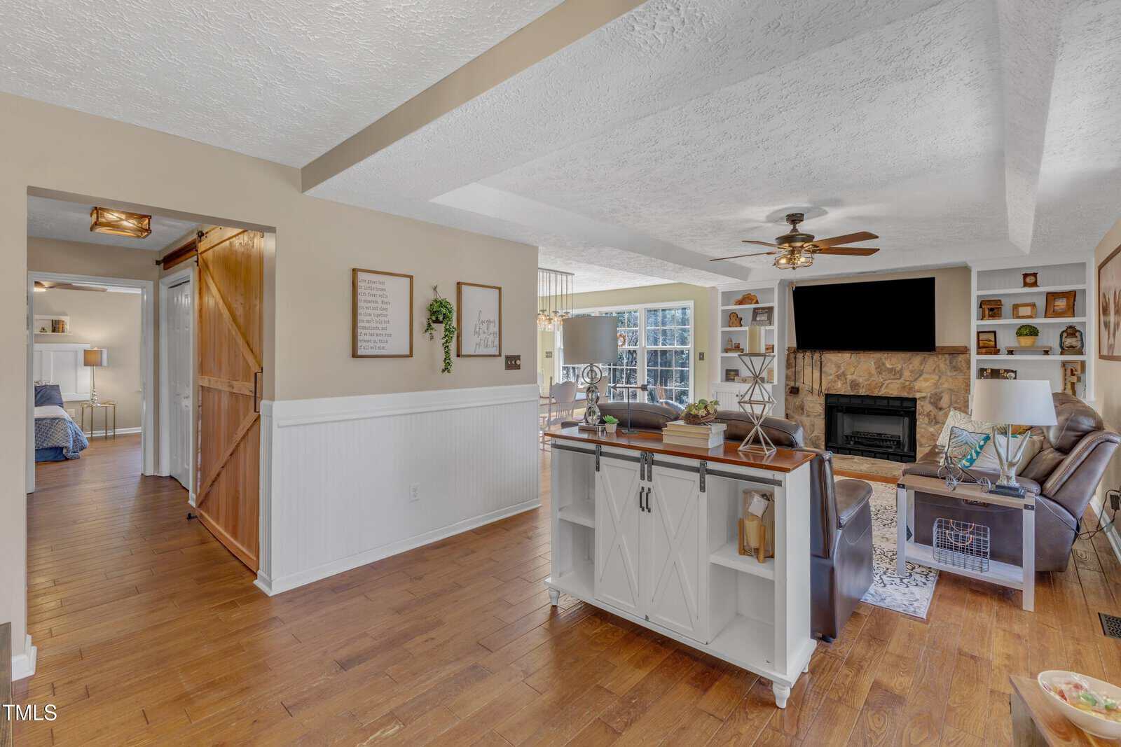 Open-concept living area with wooden flooring, a ceiling fan, and a wall-mounted TV above a stone fireplace. A kitchen island with shelves and books is in the foreground. A doorway with a sliding barn door leads to an adjacent room.