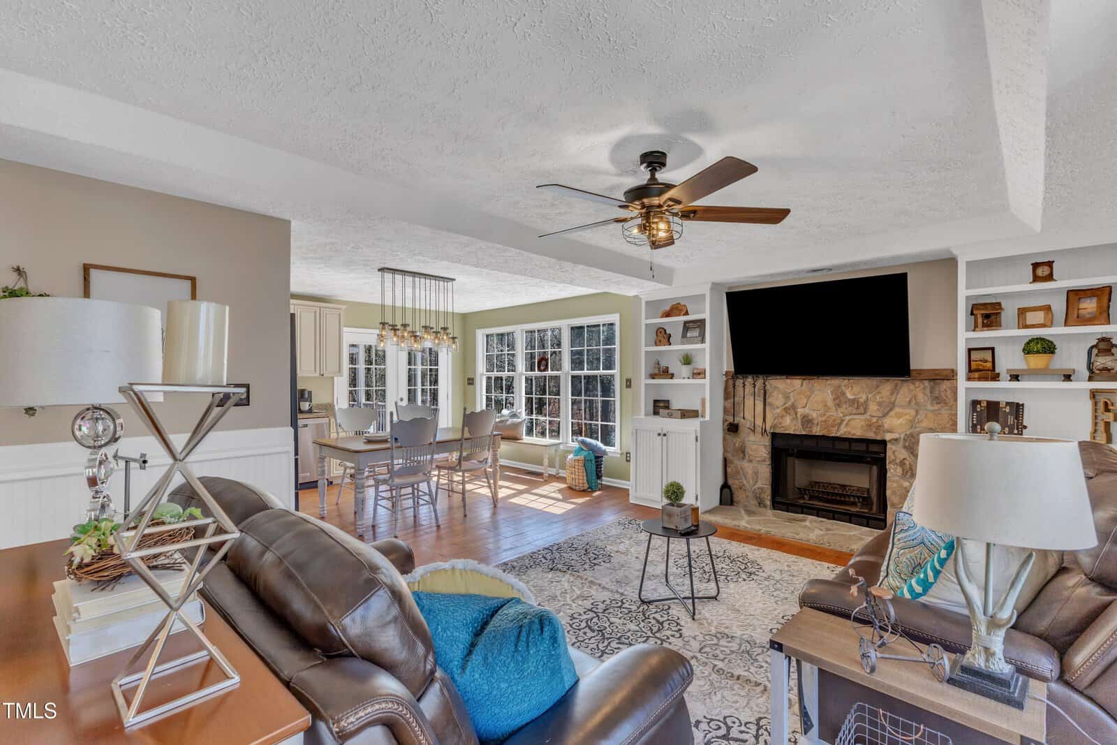 Spacious living room with leather sofas, a fireplace, and built-in shelves. A ceiling fan is mounted above. A large TV is placed over the fireplace. An adjacent dining area with a table and chairs is visible next to windows. A kitchen is in the background.