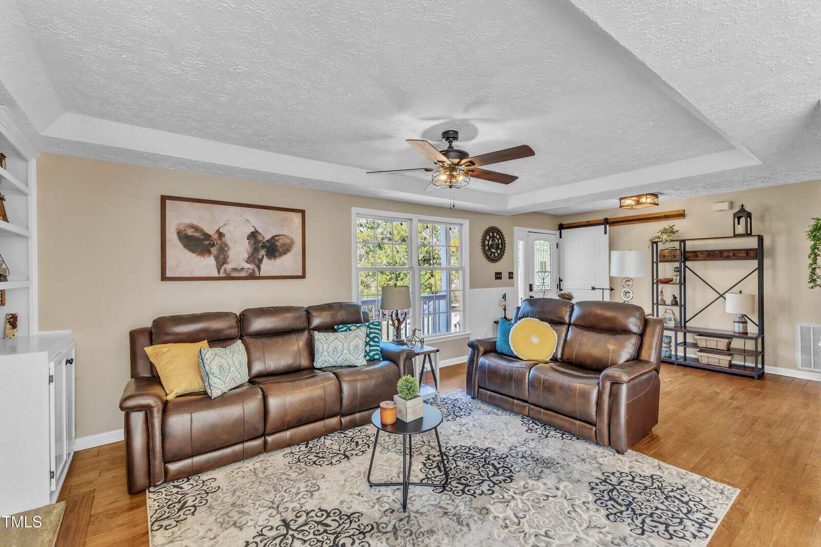Living room with two brown leather couches, a small round coffee table, and a ceiling fan. A cow painting hangs on the wall, and a sliding barn door reveals a bright room. Decorative pillows and plants add accents. A geometric-patterned rug covers the floor.