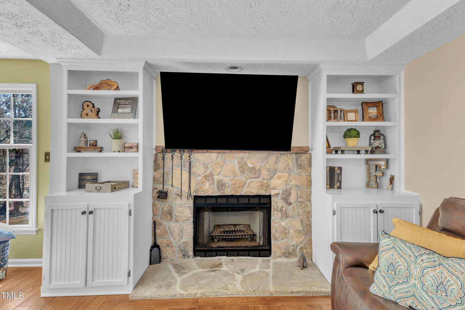 A living room features a stone fireplace with a mounted TV above. Flanking the fireplace are two white built-in bookcases displaying decor items. A window is on the left, and part of a brown couch with patterned cushions is visible on the right.