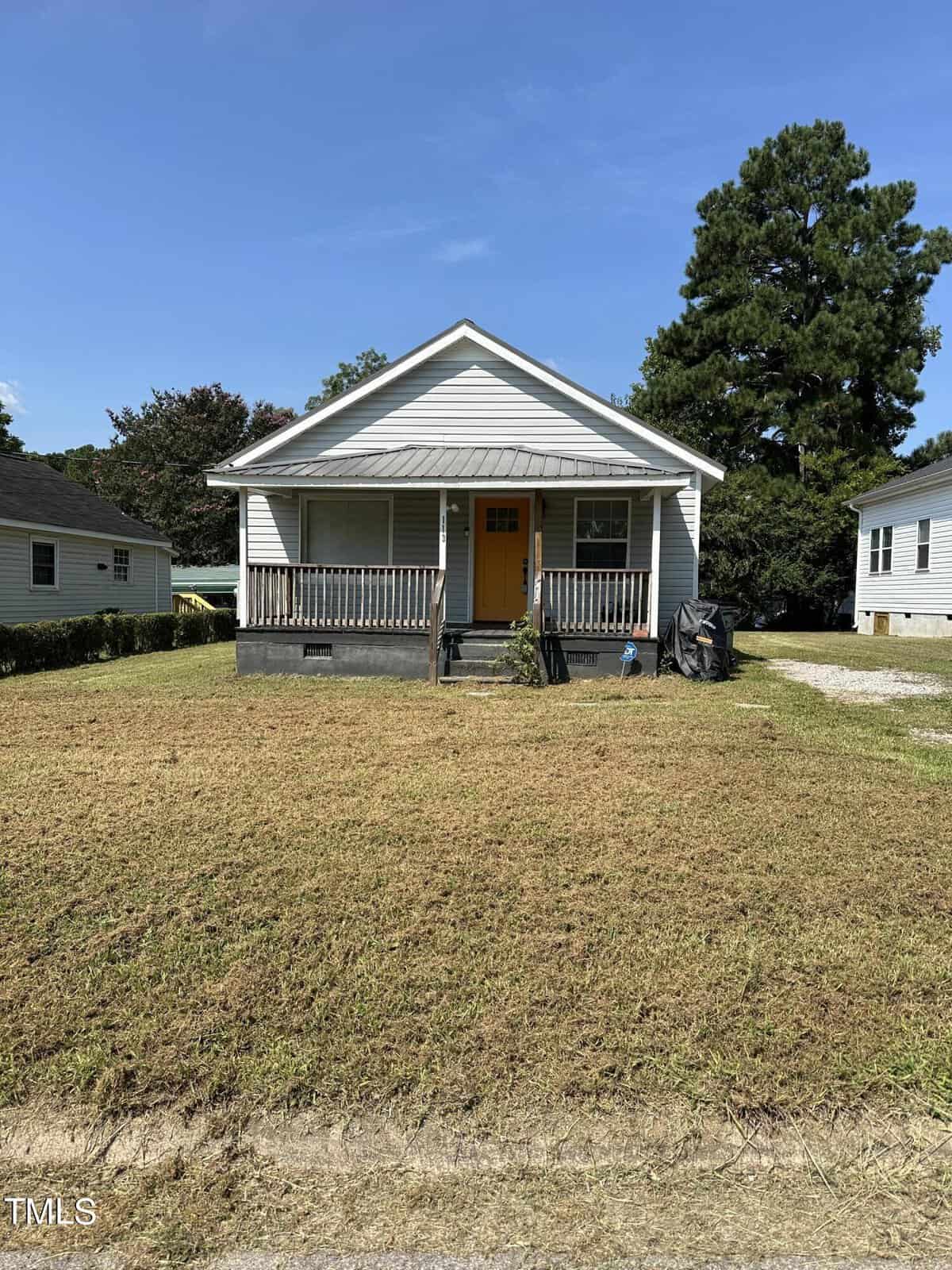 A small, single-story house at 113 Warren Avenue in Oxford boasts a gray exterior with a charming yellow front door. It features a covered porch and a freshly cut lawn. A large tree stands tall in the background, flanked by neighboring houses under the clear, sunny sky.