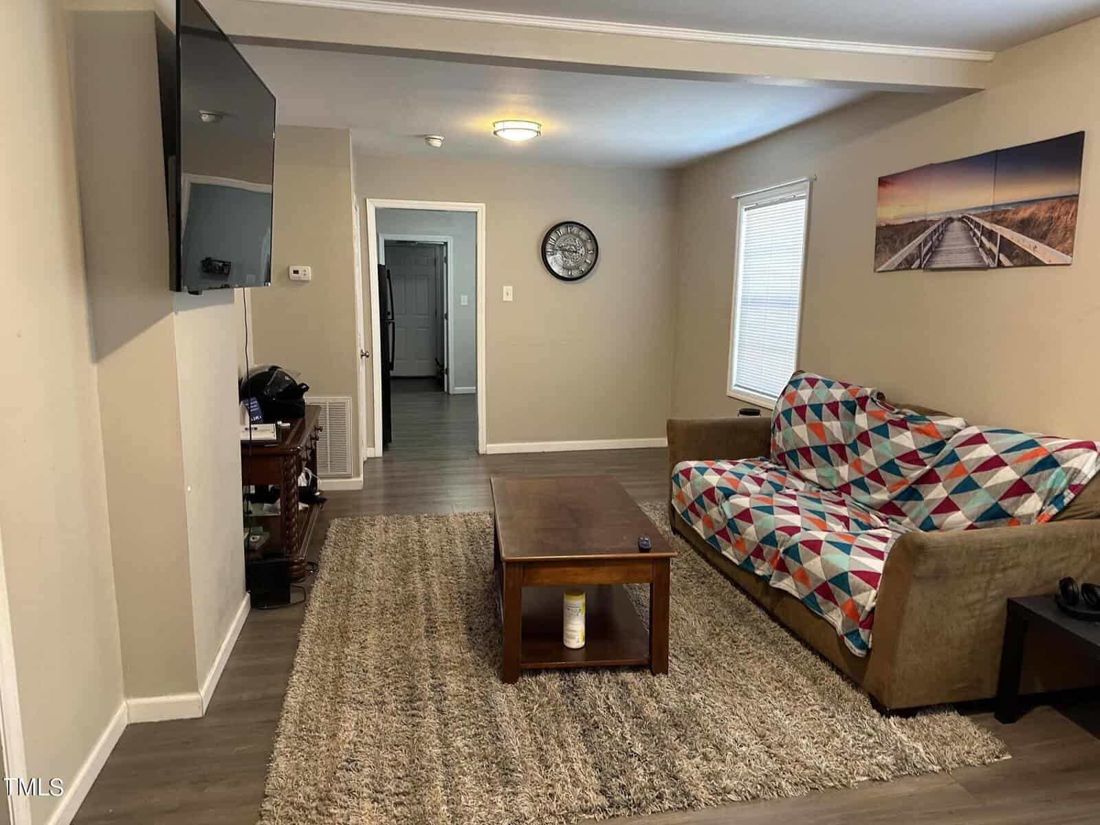 Living room with a wall-mounted TV, a wooden coffee table, and a sofa covered with a multicolored quilt. The room features a neutral color scheme, a shaggy area rug, and a landscape picture on the wall. There's a doorway leading to another room.