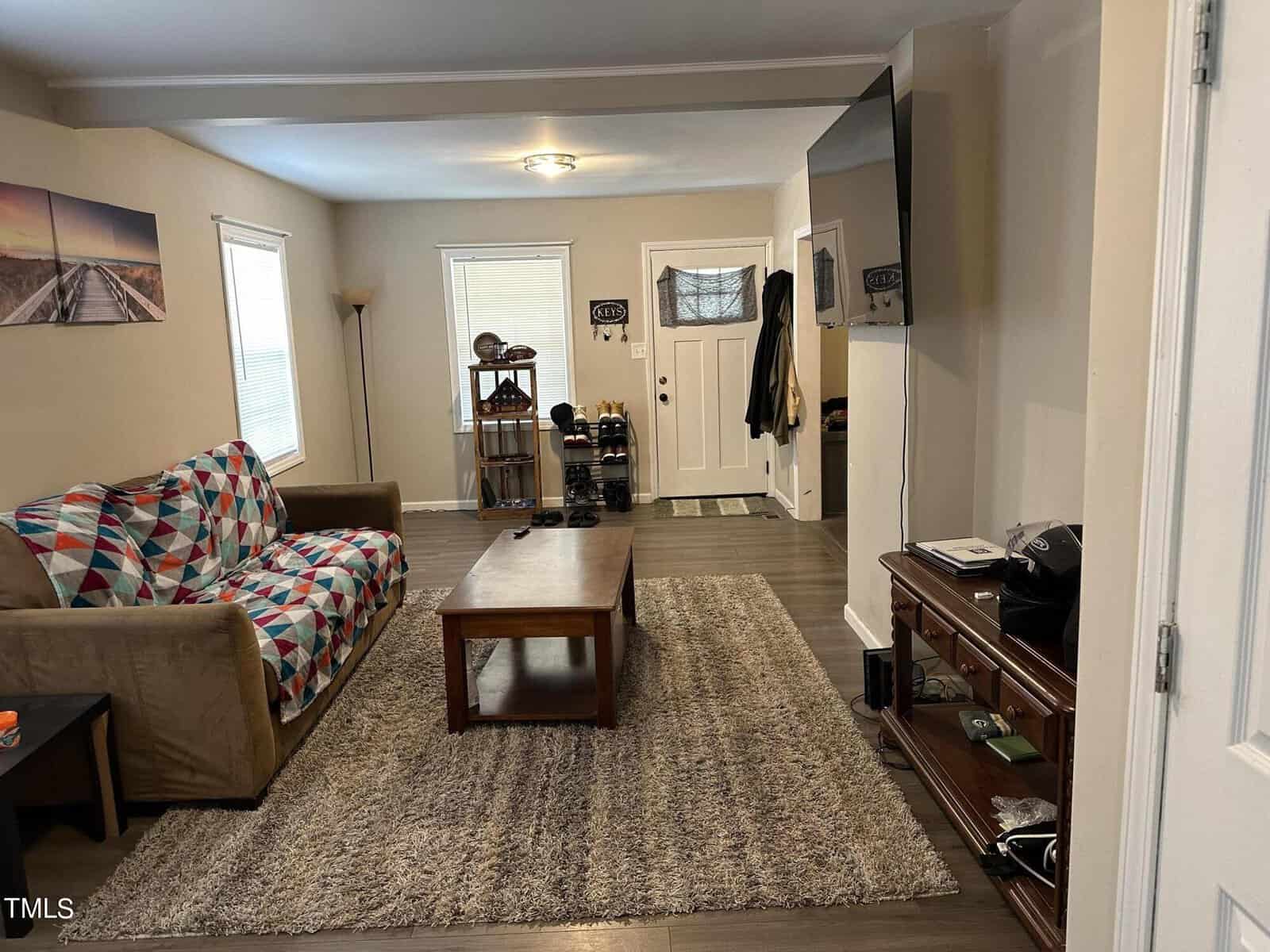 Living room with a gray rug on wooden flooring, a brown couch with a colorful quilt, a wooden coffee table, and a TV on the wall. There's a shoe rack by the front door and framed artwork on the walls.