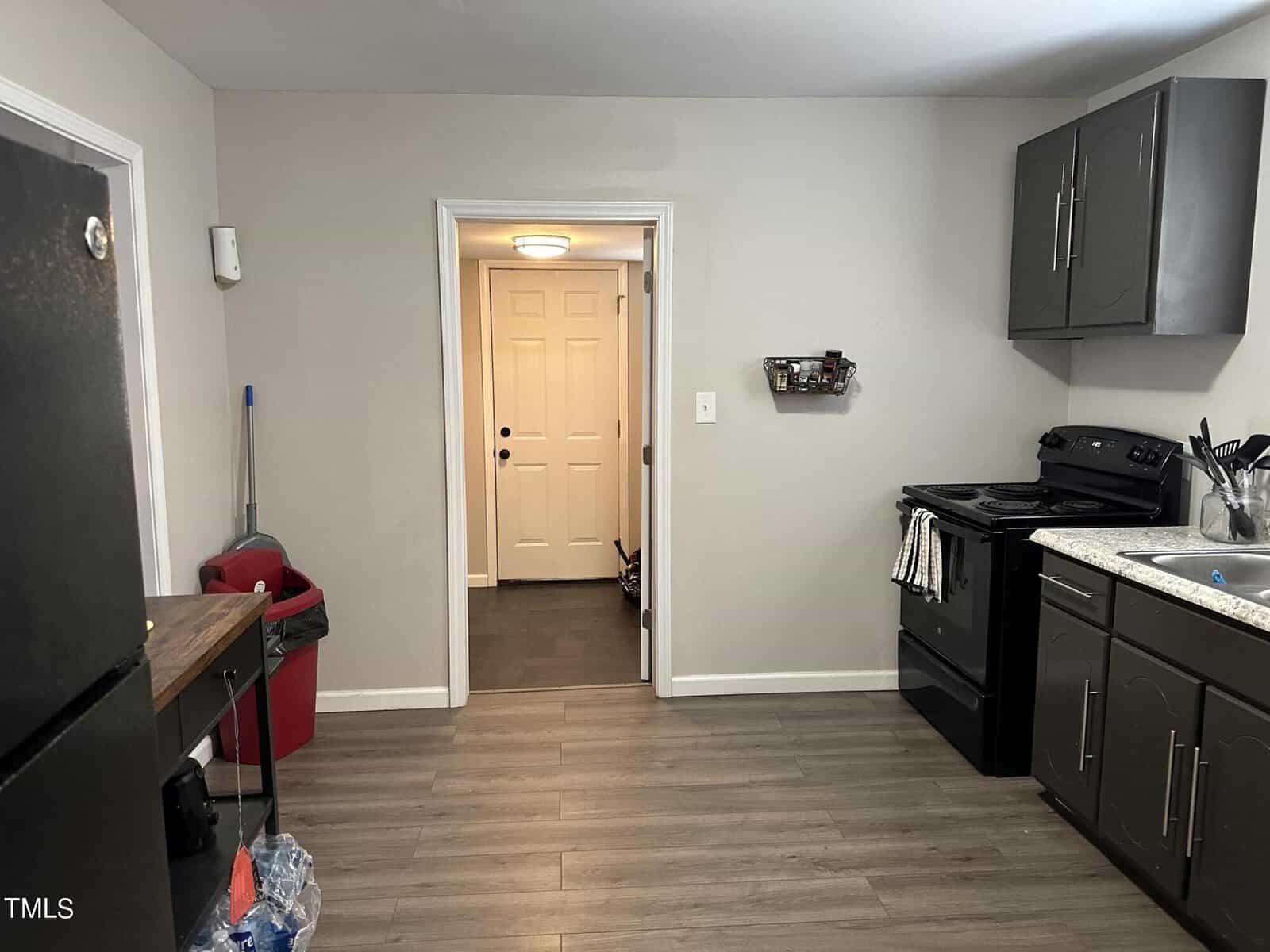 A kitchen with gray cabinetry and wood floors. There's a black stove and refrigerator, with a sink and countertop to the right. A doorway leads to a hallway with a closed white front door. Household items are visible in the corner.