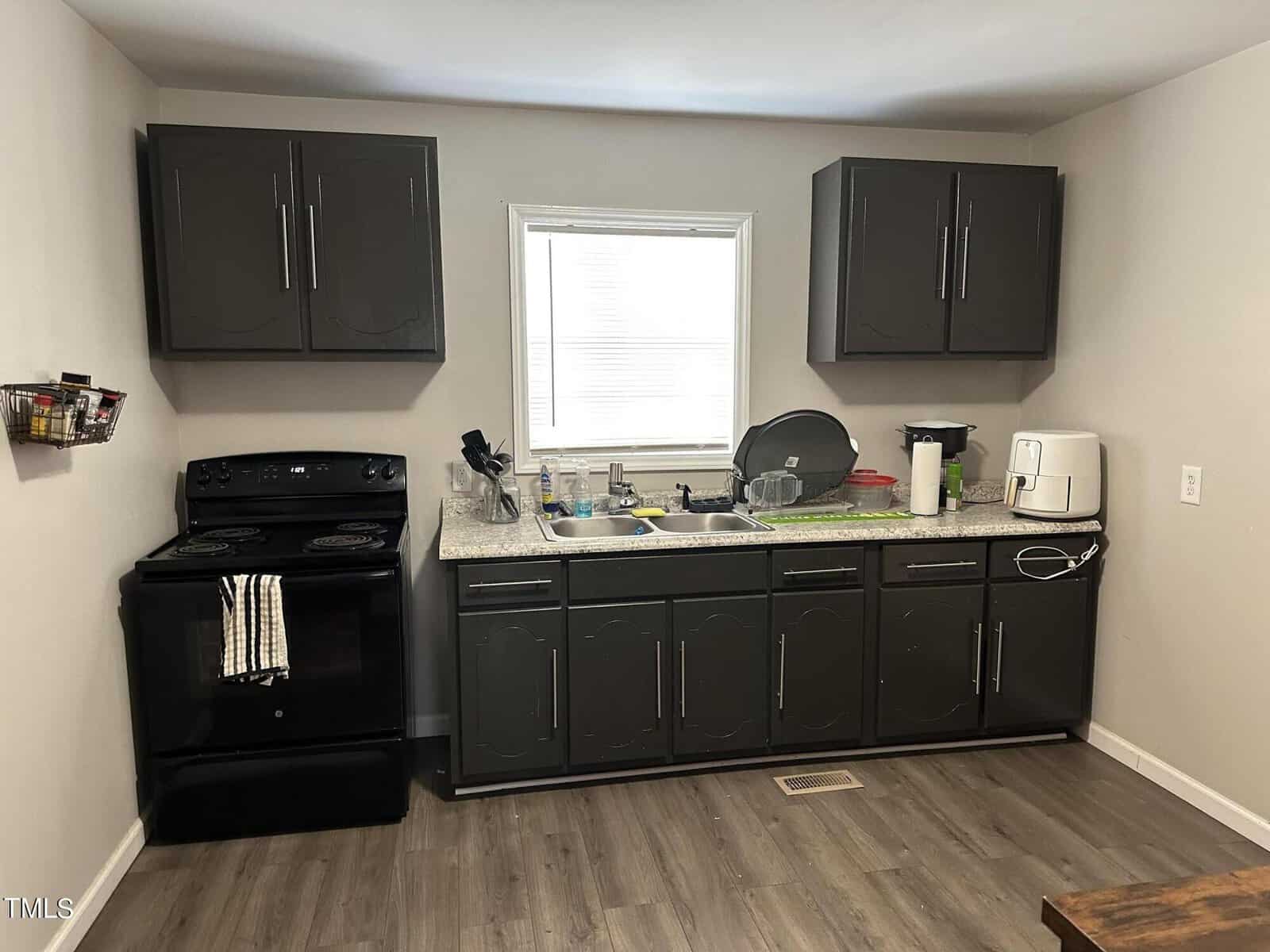 A compact kitchen with a black stove, gray cabinets, and laminate flooring. The countertop has a sink, drying rack with dishes, and a coffee maker. A window with blinds is above the sink. A towel hangs on the stove handle.