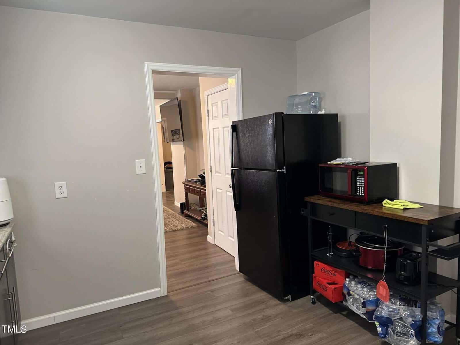 Kitchen area with a black refrigerator and a black microwave on a wooden cart. The cart holds various kitchen items and bottled water. Gray walls and wood flooring are visible. An open door leads to a hallway with a glimpse of a room beyond.