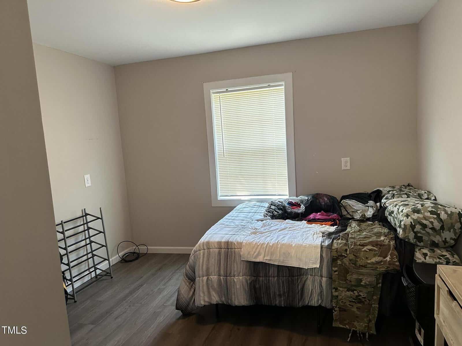 A sparsely furnished bedroom with a bed covered in mixed patterned coverings, including camouflage. Clothes are piled on the bed. There's a small metal shoe rack on the left, and a window with closed blinds above the bed. The floor is wooden.