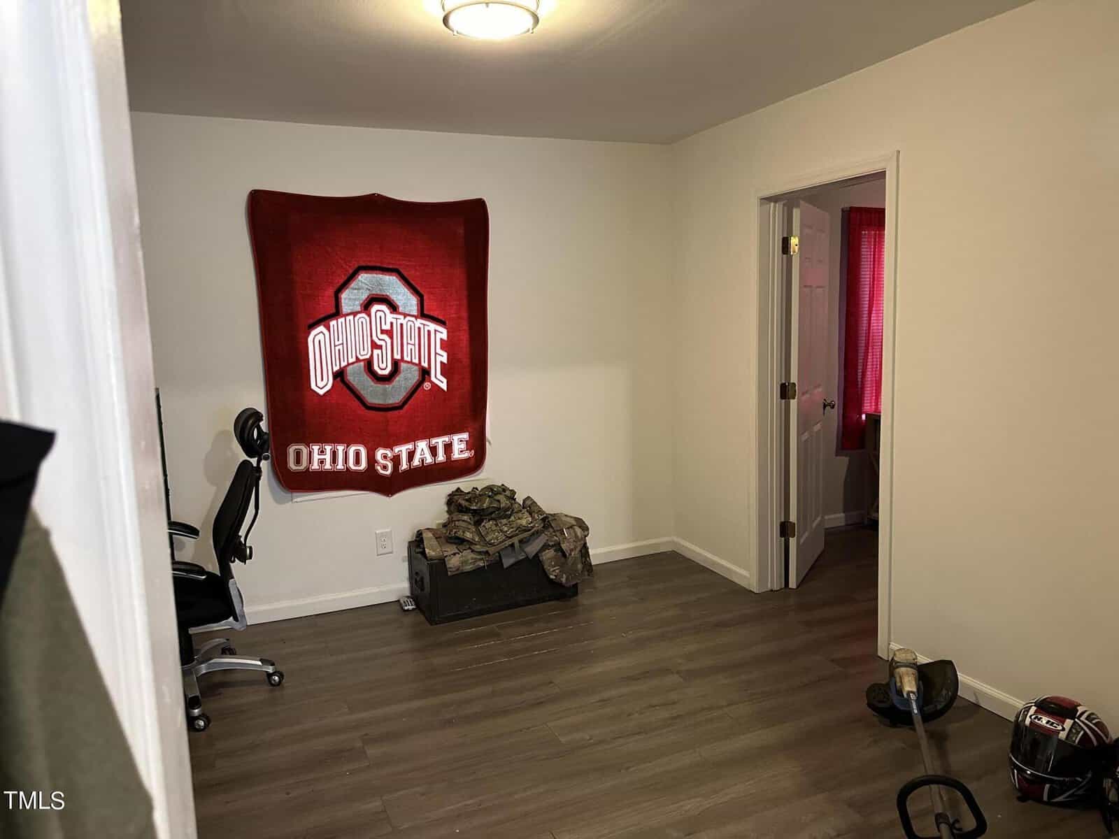 A small room with a wooden floor features an Ohio State blanket hanging on the wall. There is an office chair in the corner, a box covered with a blanket, and a football helmet on the floor. A doorway leads to another room.