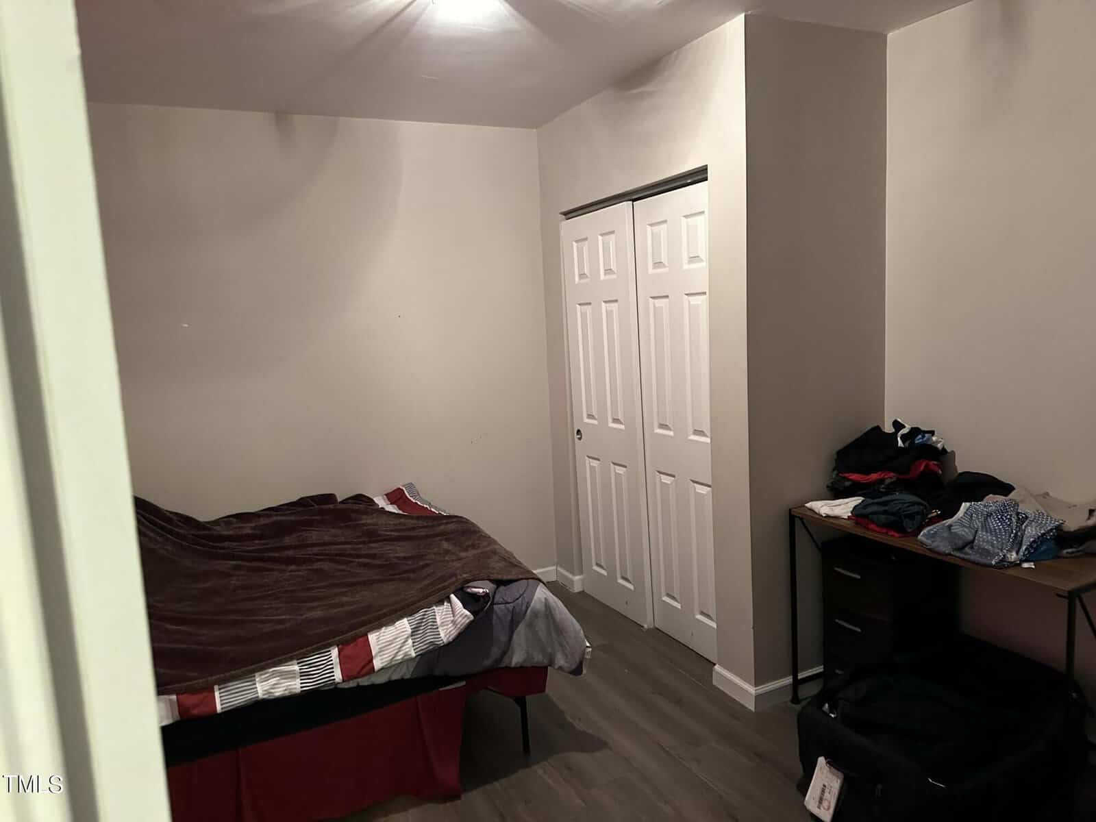 A small bedroom with beige walls features a bed with a brown blanket and striped sheets. There's a closet with white sliding doors and a black desk with clothes piled on top. The room has wood flooring.